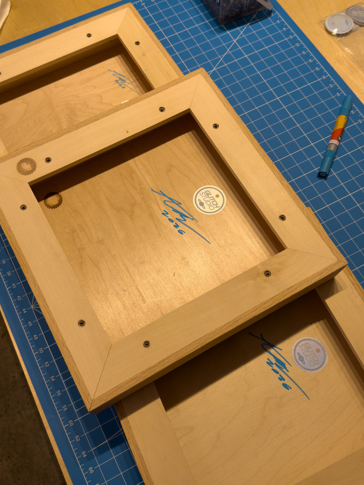 Three unfinished light wood picture frames laid out on a blue grid cutting mat, with visible screw holes and a small round sticker inside each frame.