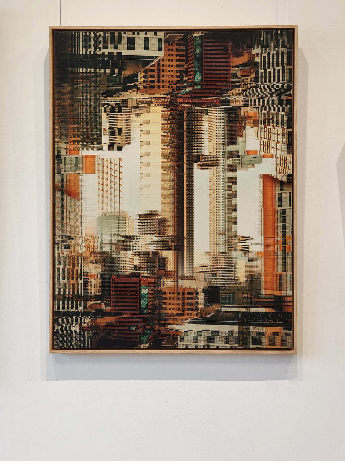 A framed abstract cityscape artwork featuring overlapping skyscrapers in warm brown, beige, white, and orange tones, hung on a white wall.