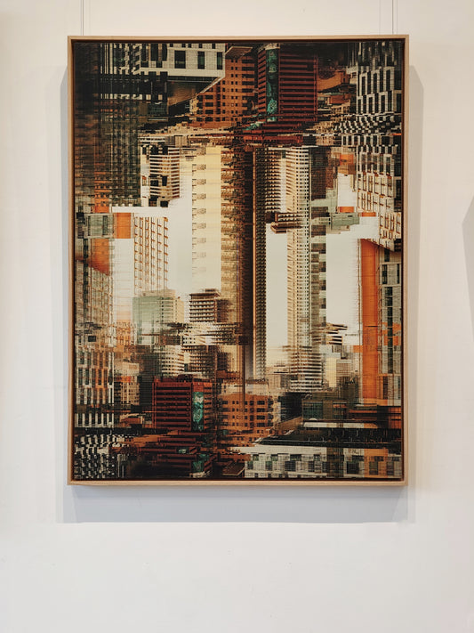 A framed abstract cityscape artwork featuring overlapping skyscrapers in warm brown, beige, white, and orange tones, hung on a white wall.