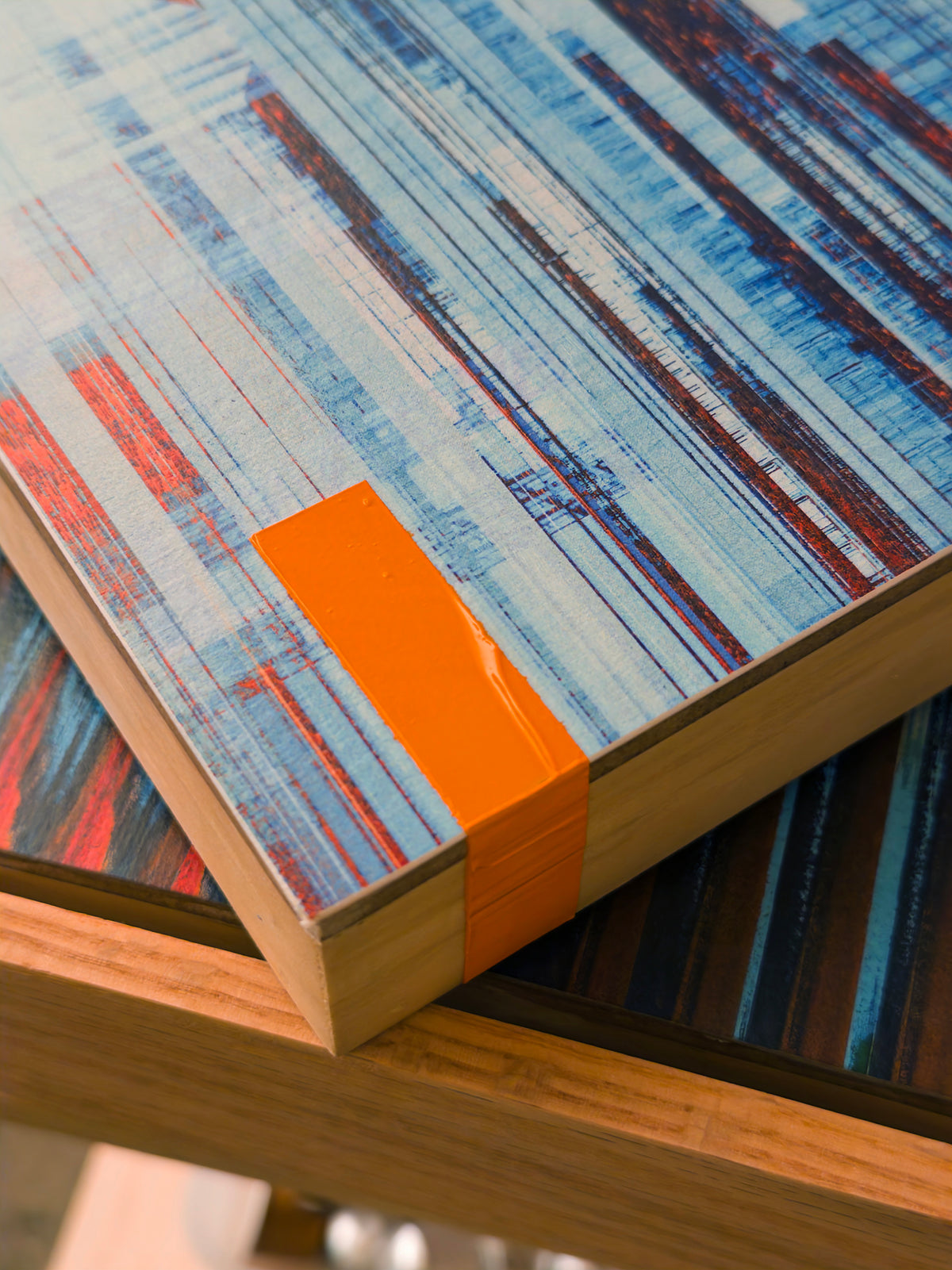 Close-up of a rectangular wooden board or tray with an abstract painted top featuring blue, white, red, and dark brown brushstrokes, with a bright orange rectangular accent on the side.