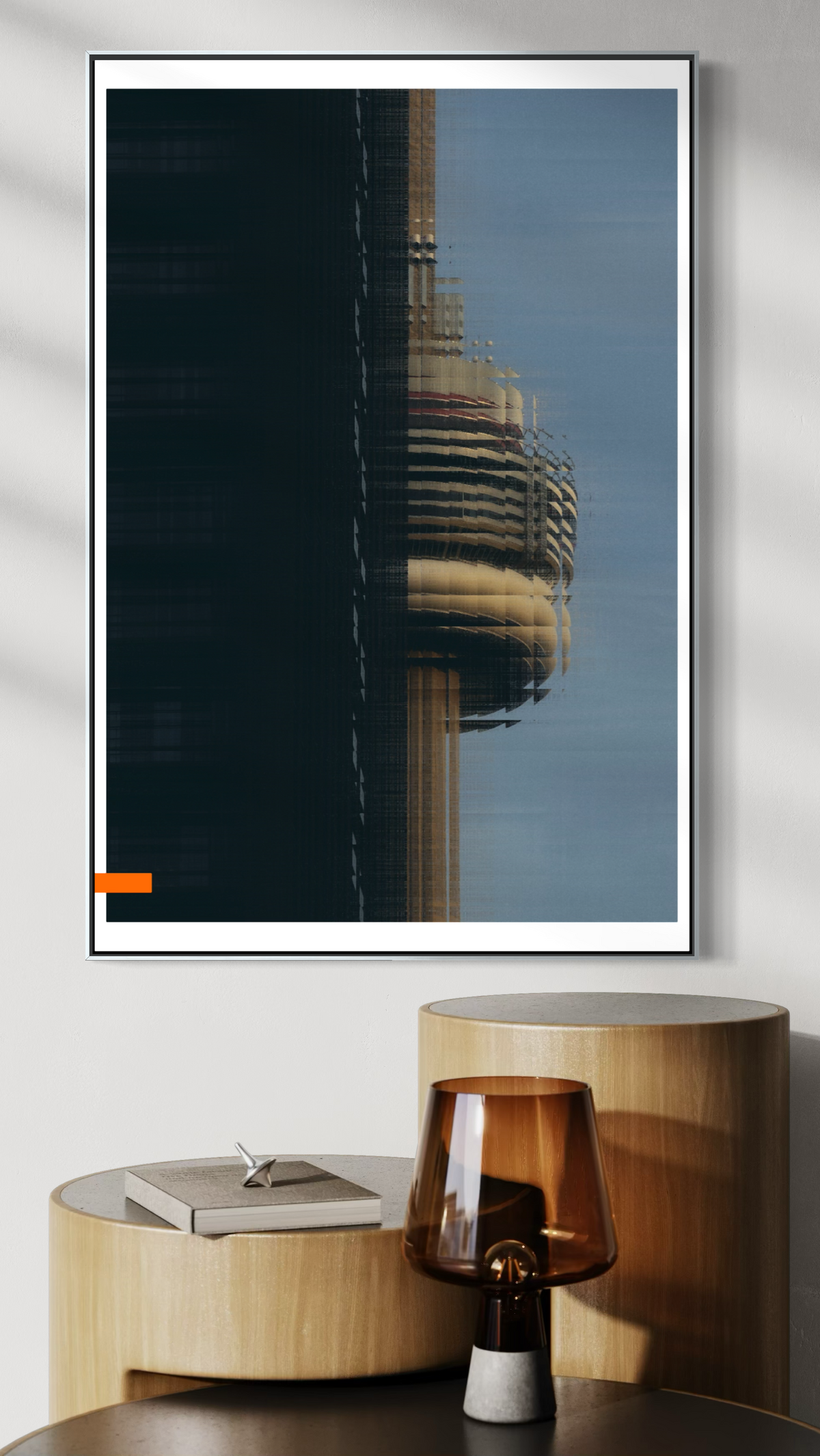 A framed vertical art print featuring a modern skyscraper with the CN tower, displayed on a light gray wall above a wooden side table with a lamp and books.