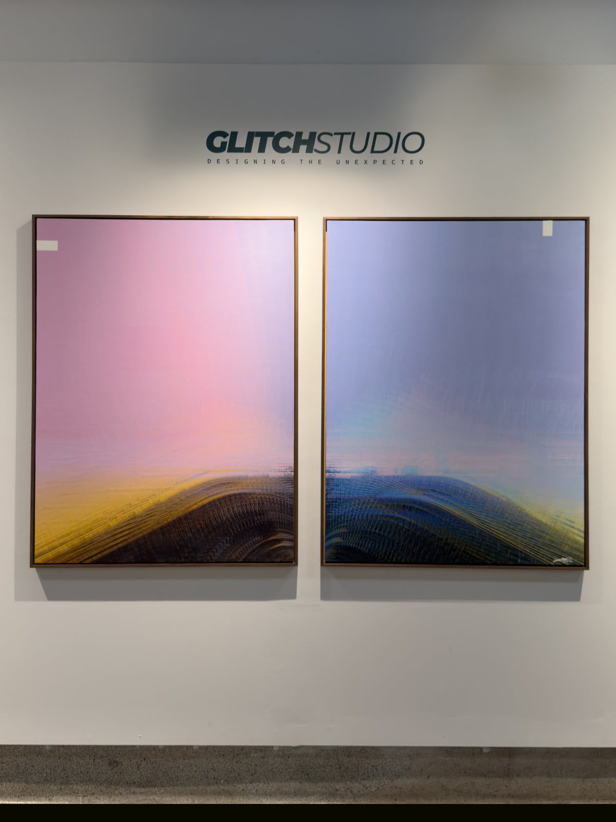 Two framed abstract landscape art prints displayed side-by-side on a white gallery wall under the brand name GLITCHSTUDIO. The left print features a pink-to-yellow gradient over dark forms, and the right print shows a blue-to-yellow gradient over dark forms.