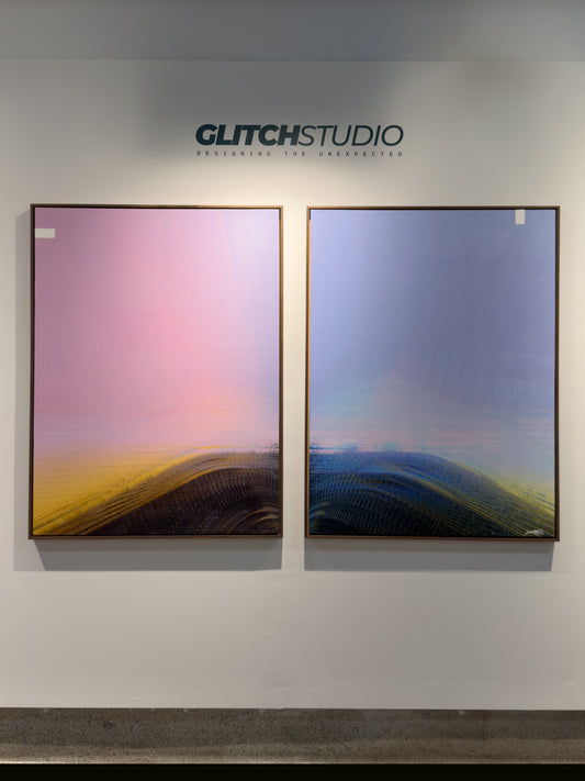 Two framed abstract landscape art prints displayed side-by-side on a white gallery wall under the brand name GLITCHSTUDIO. The left print features a pink-to-yellow gradient over dark forms, and the right print shows a blue-to-yellow gradient over dark forms.